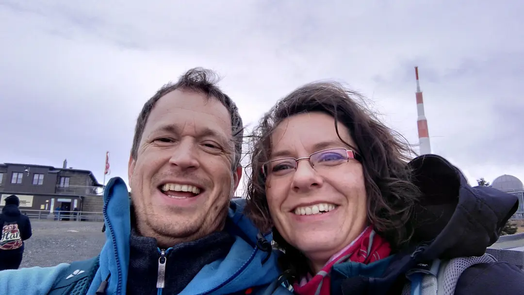 Selfie of Manu & Markus with tousled hair in front of the transmitter antenna on the Brocken