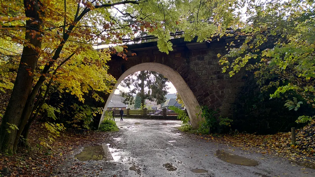 Ein steinerner Brückenbogen unter dem die ersten Gartenzäune eines Ortes sichtbar sind