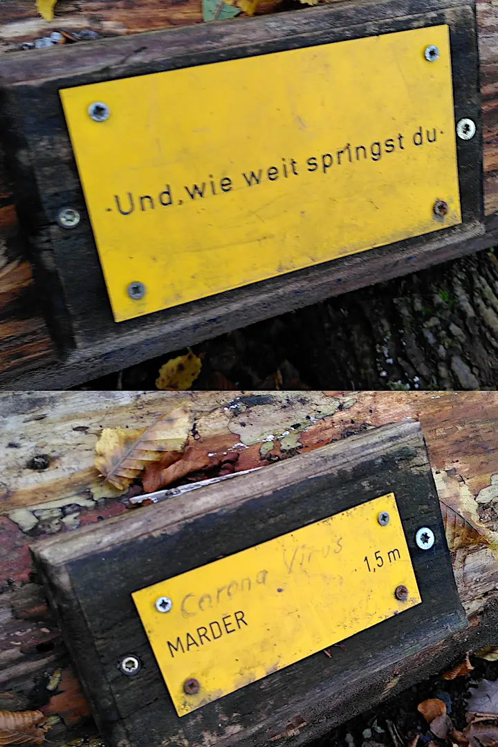 Zwei Schilder mit der Aufschrift "Und wie weit springst du?" und "Marder 1,5 m". Jemand hat auf das Marder-Schild mit Kuli "Corona-Virus" geschrieben