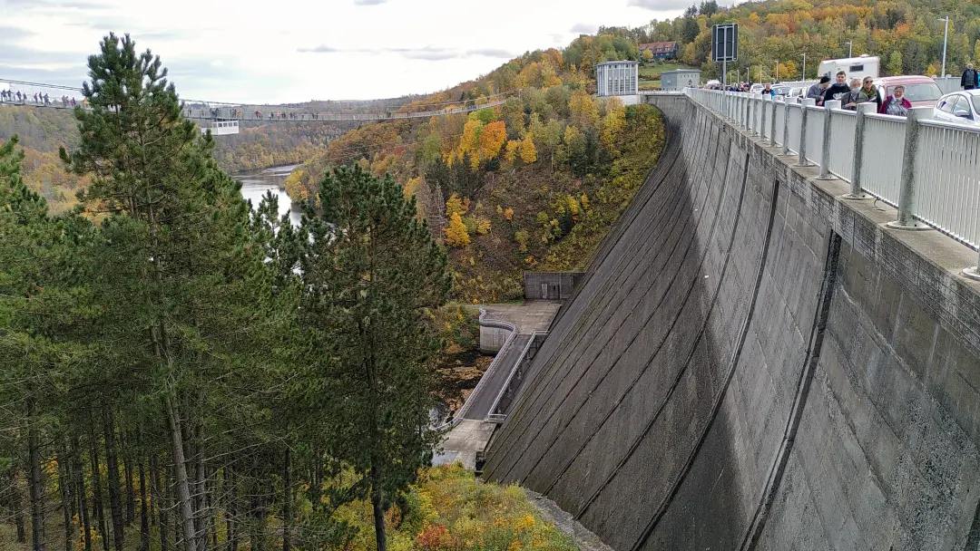 Blick entlang der Krone der Staumauer der Rappbode-Talsperre. Über dem Tal ist eine Hängebrücke für Fußgänger zu erkennen, die den Auslauf der Staumauer überspannt.