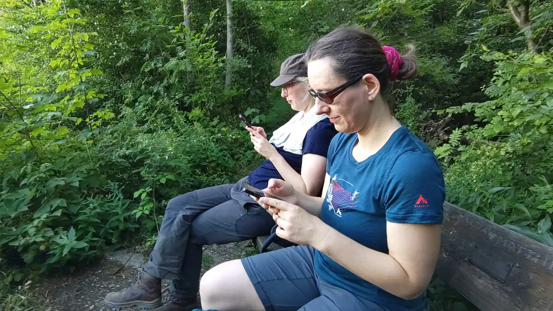 Lütti und Manu sitzen auf einer Bank im Wald und schauen in ihre Handys