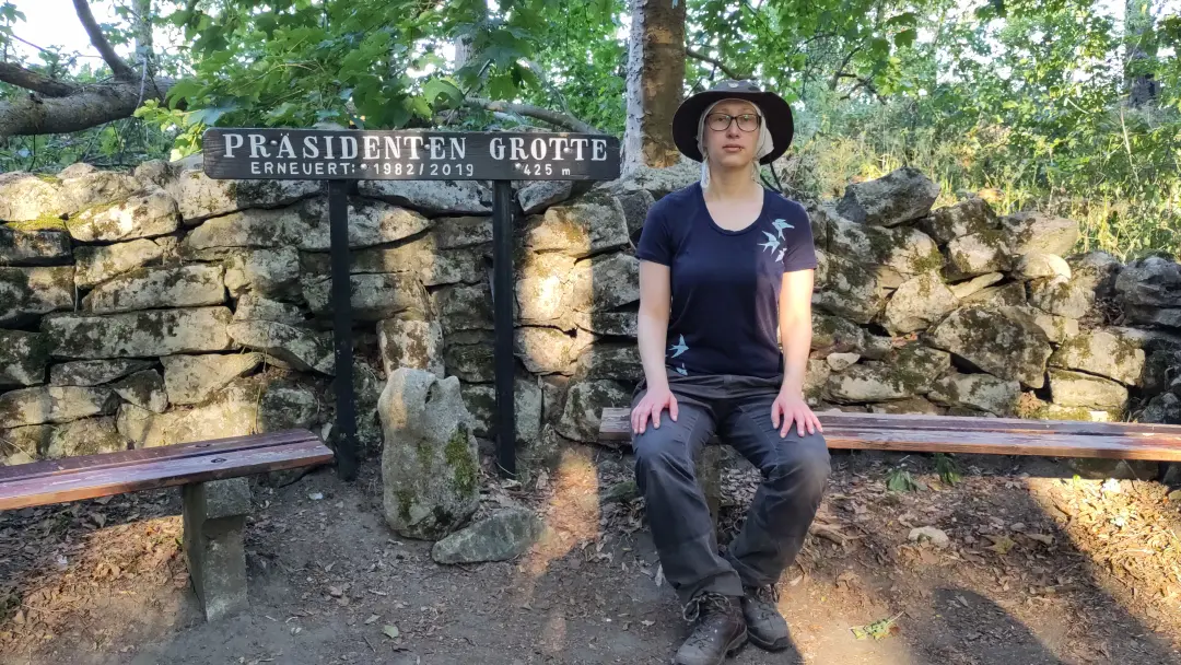 Lütti sitzt auf einer Bank vor eine Trockenmauer mit dem Schild "Präsidenten-Grotte Erneuert 1982/2019 425 m"