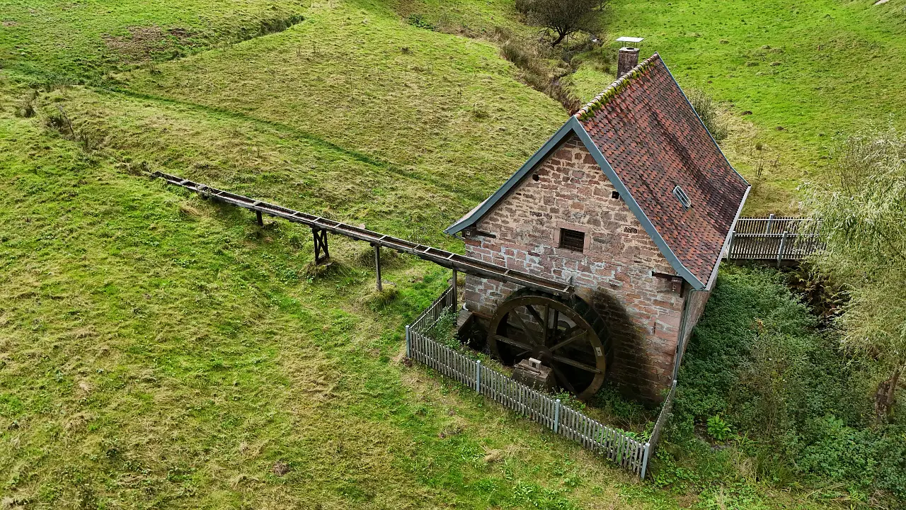 The old village mill in Habichtstal seen from the air. You can see the old stone building with the waterwheel on the side standing in a green meadow.