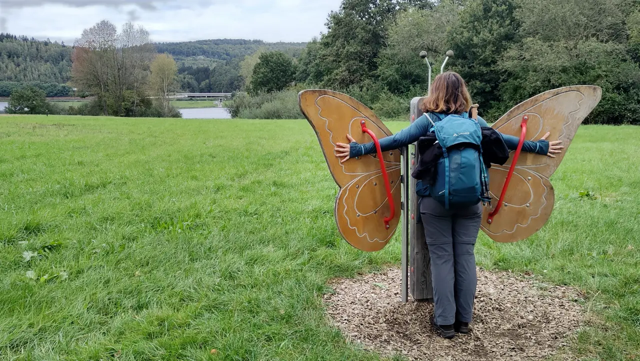 Manu sticking her arms through the large wooden wings of a butterfly sculpture by the path. The artwork is designed so that people form the butterfly's body and "wear" the wings while enjoying the view toward the Kinzigtalsperre.