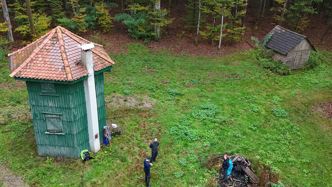 Aerial view of Waidmannsruh, a hexagonal green tower with a red tile roof in the middle of the forest. At the edge of the picture you can see a large fire pit.